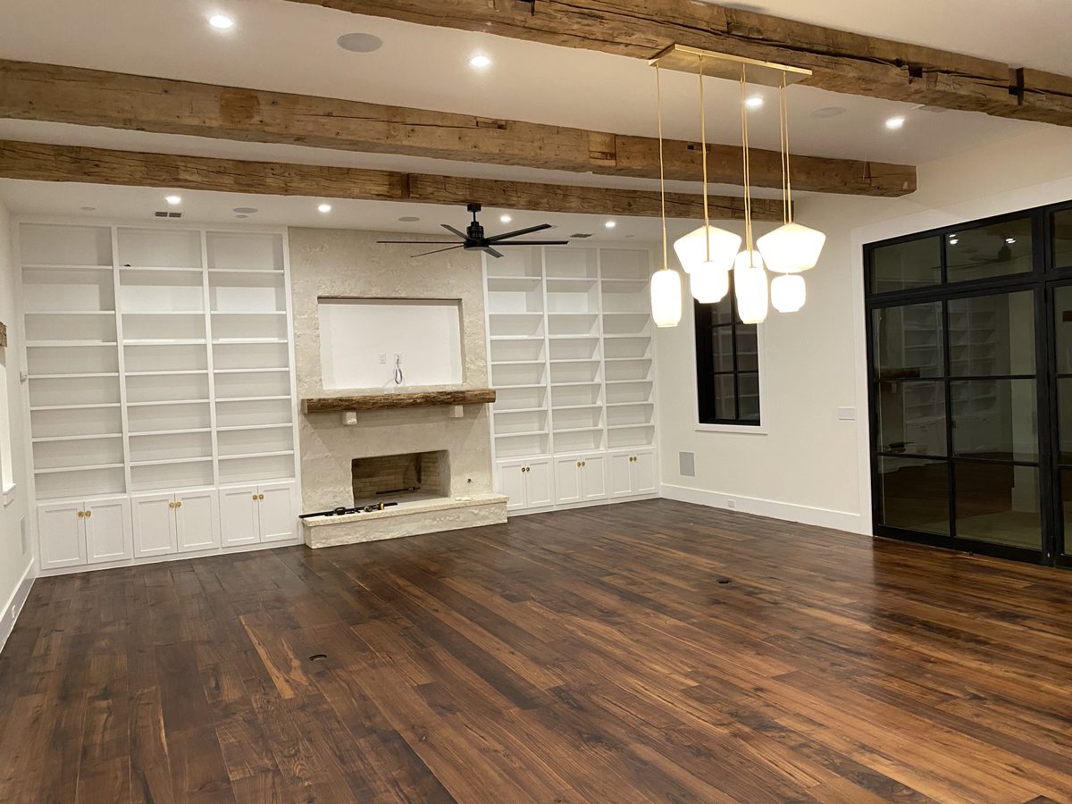 Floor-to-ceiling built-in bookcases with cabinet bases flanking a limestone fireplace, under a ceiling of exposed reclaimed beams.