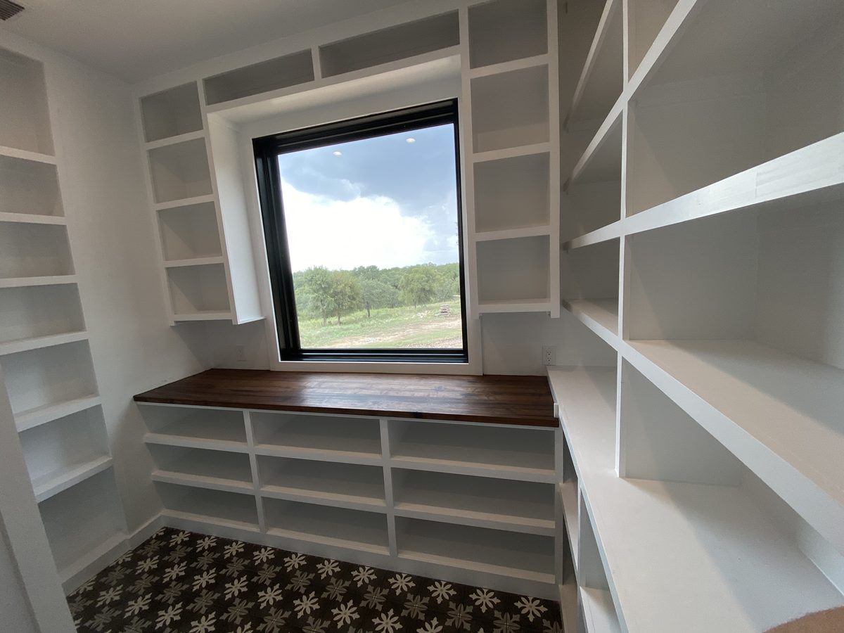 Walk-in pantry lined with custom white shelving on three walls and a stained solid wood counter beneath a black window.
